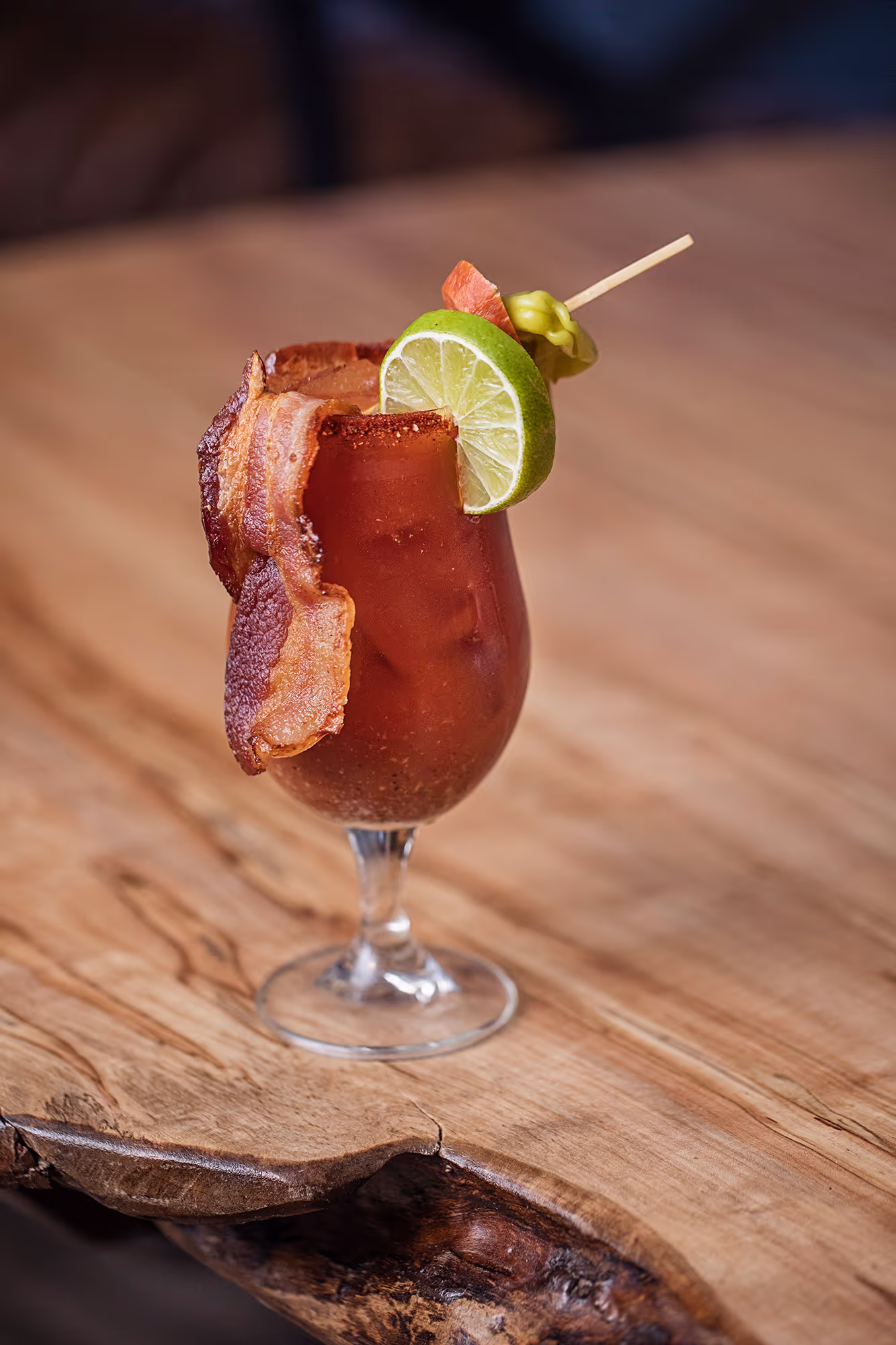 A garnished cocktail with bacon, lime, and tomato, served in a glass on a rustic wooden table.