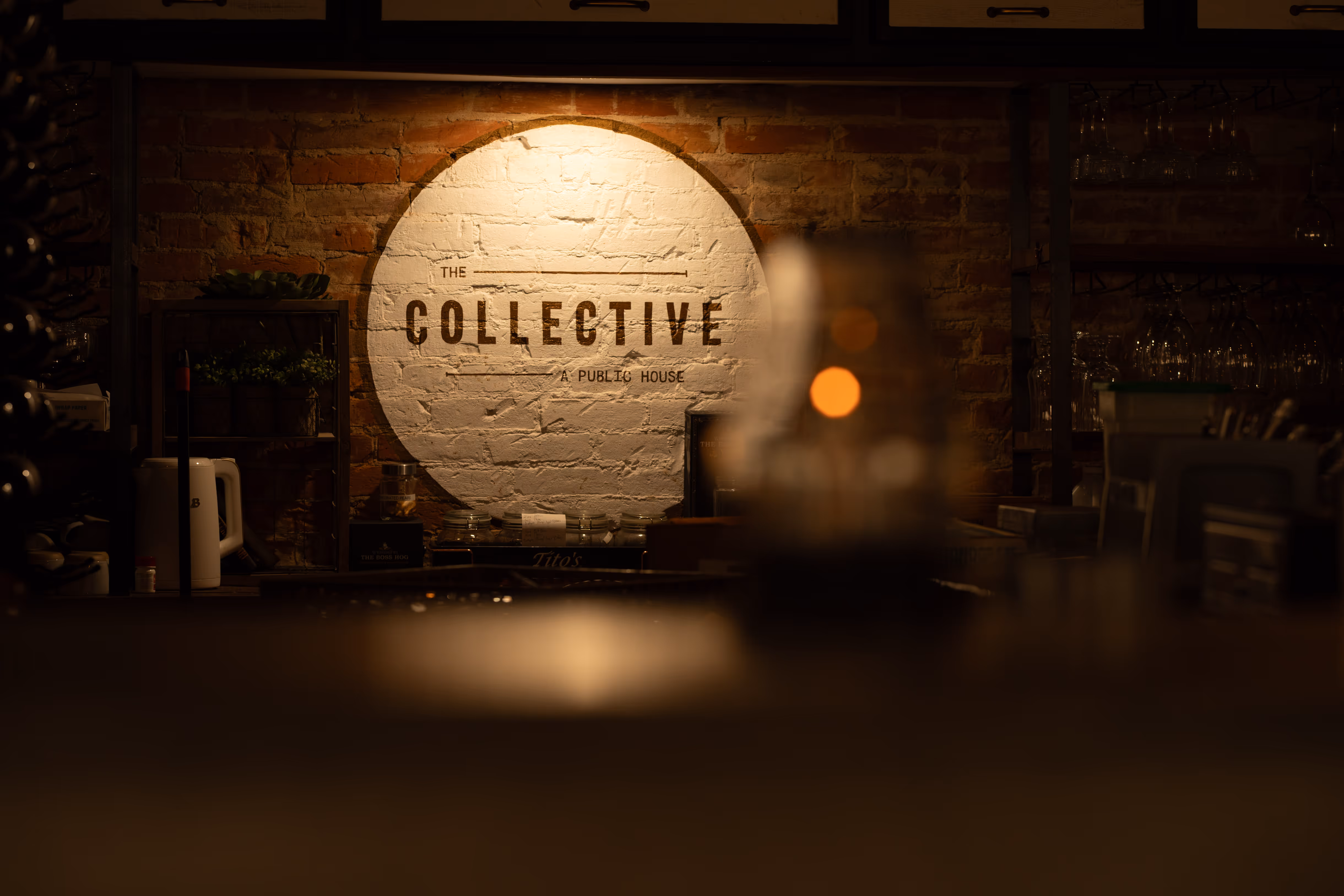 Dimly lit interior with a round sign reading "THE COLLECTIVE A PUBLIC HOUSE" against a brick wall, showcasing a cozy bar atmosphere.