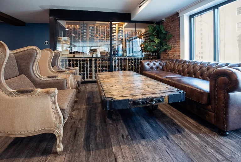 A cozy lounge featuring upholstered armchairs, a leather sofa, and a rustic wooden coffee table, with large windows and a wine rack backdrop.