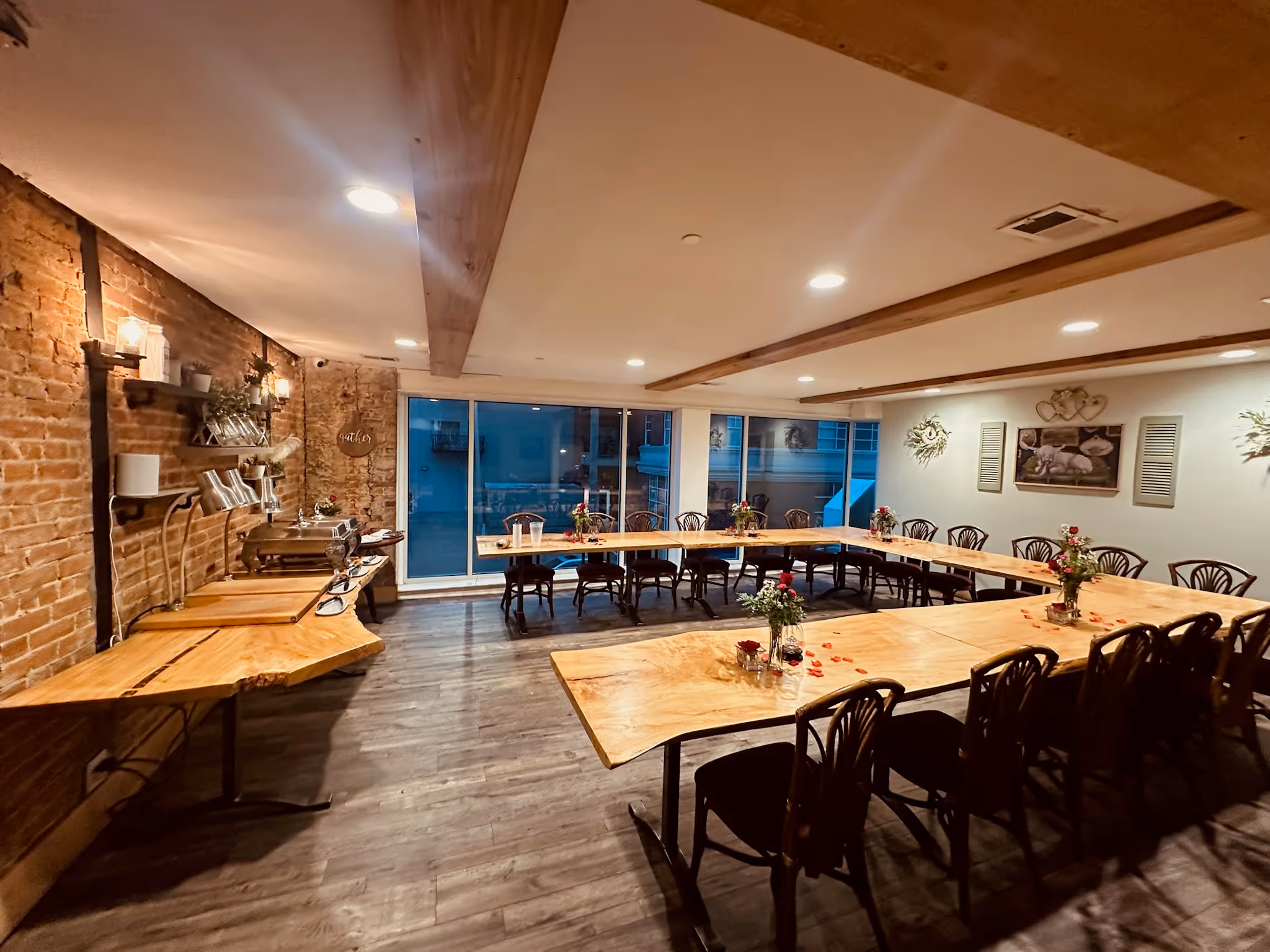 A cozy meeting room featuring wooden tables, black chairs, floral decor, and large windows, illuminated with soft lighting.