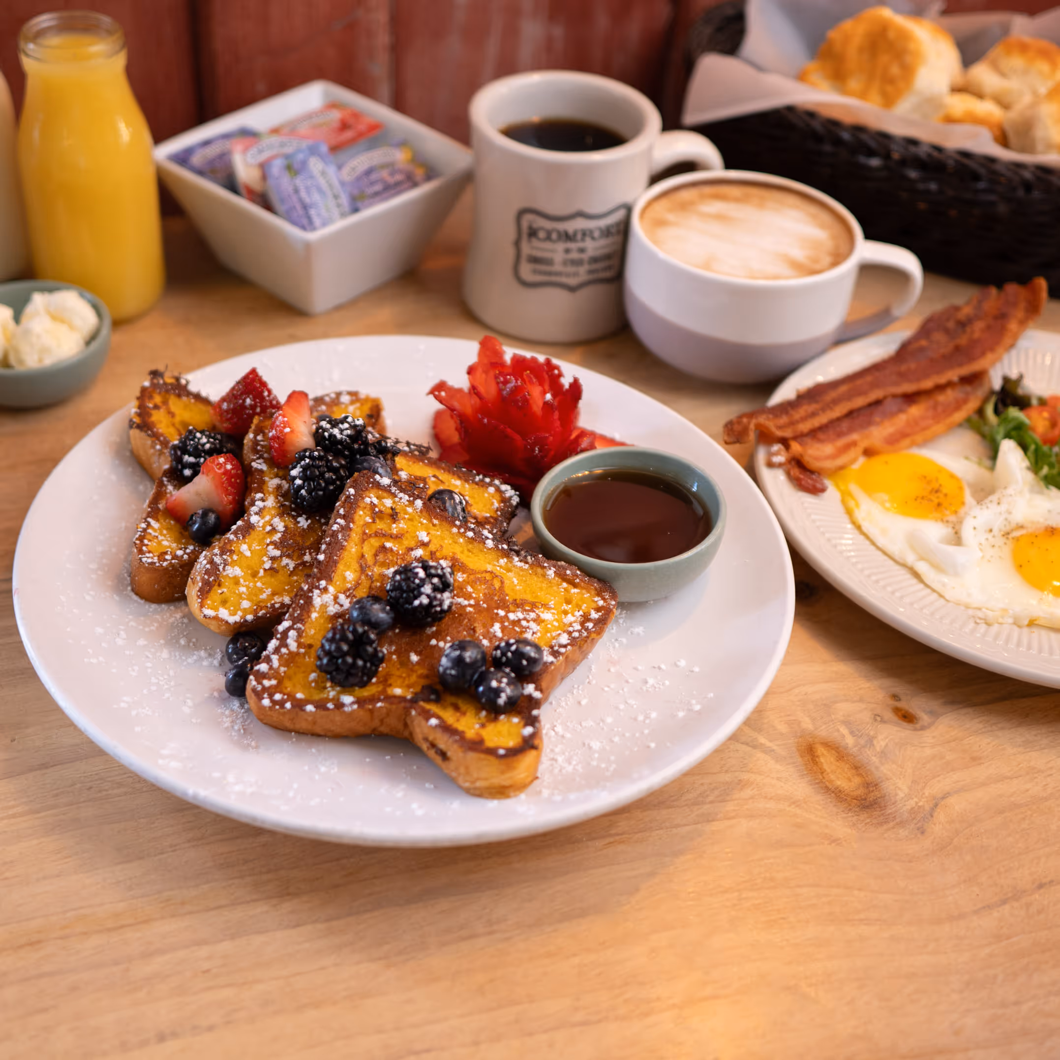A delicious breakfast spread featuring French toast with berries and syrup, eggs with bacon, juice, coffee, and biscuits on a wooden table.