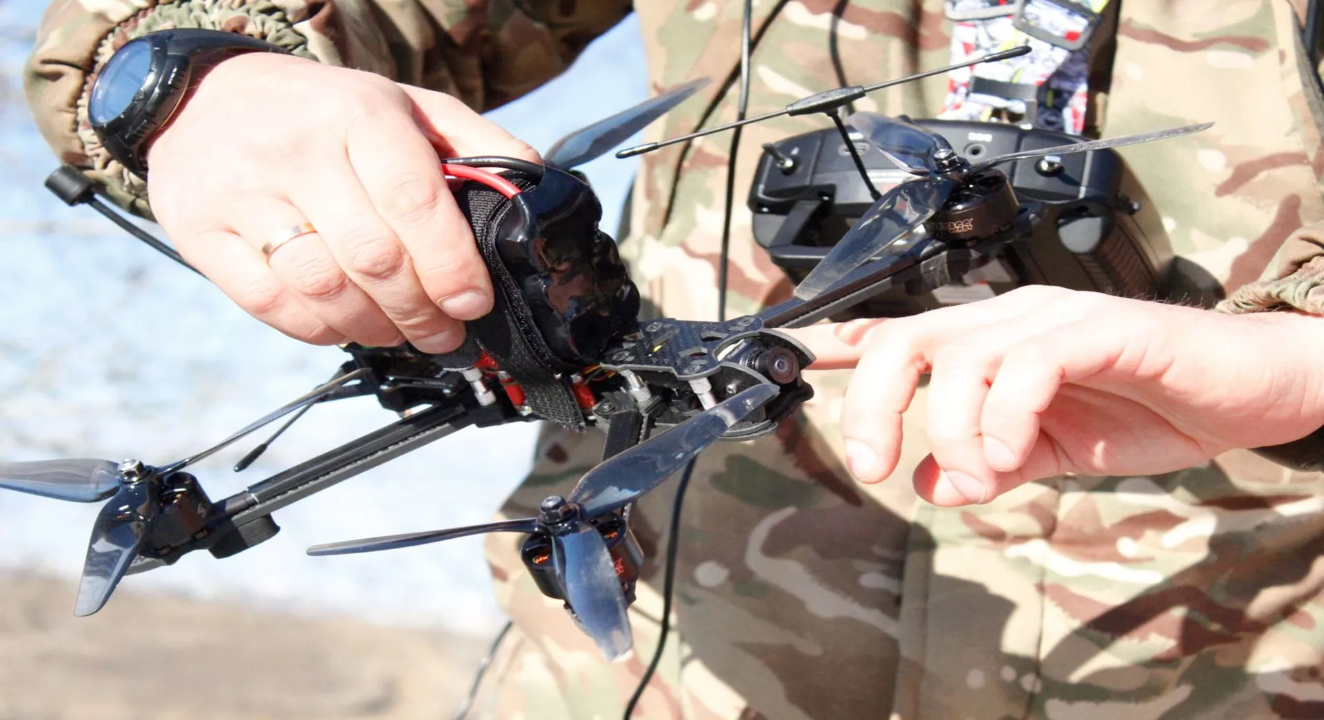 A man in camouflage clothing holds a quadcopter with four propellers and a remote control.