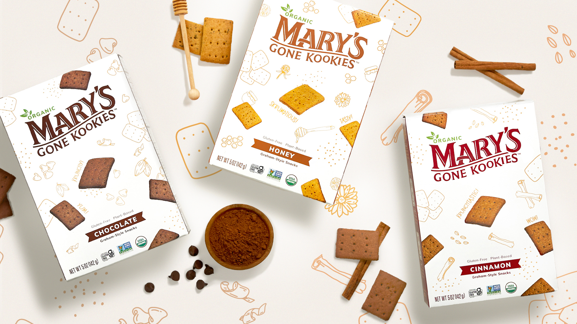 Three boxes of Mary’s Gone Kookies in different flavors surrounded by kookies, cinnamon sticks, chocolate chips, and a honey stirrer.