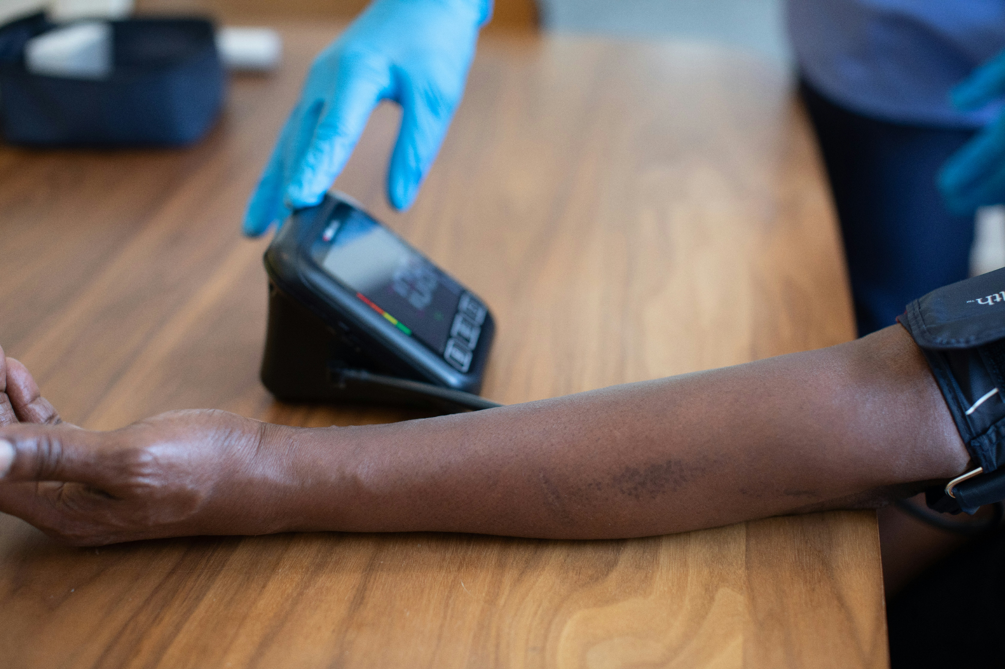 A clinician wearing gloves measures a patient’s blood pressure using an arm cuff and digital monitor on a wooden table.