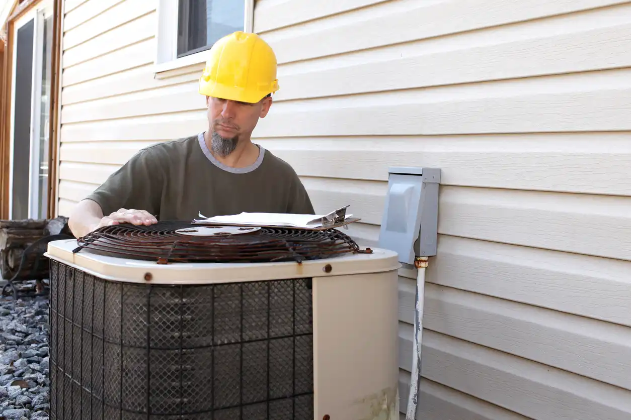 Technician in yellow hard hat and green shirt servicing outdoor AC unit beside beige house