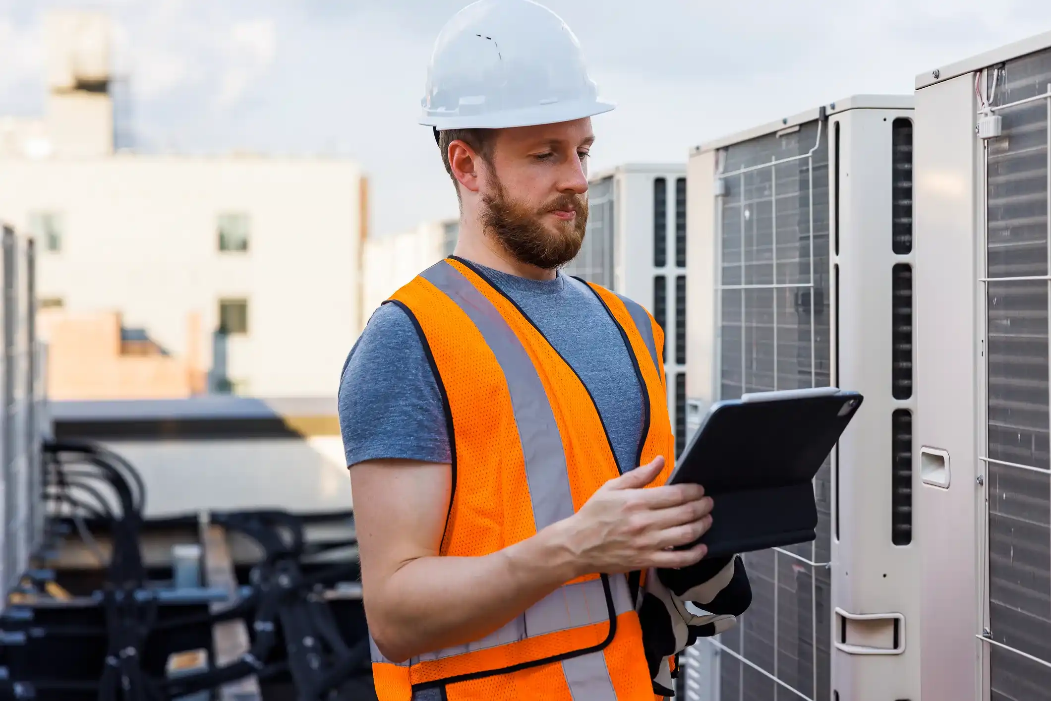 Worker in orange safety vest and white hard hat using tablet near large outdoor AC units