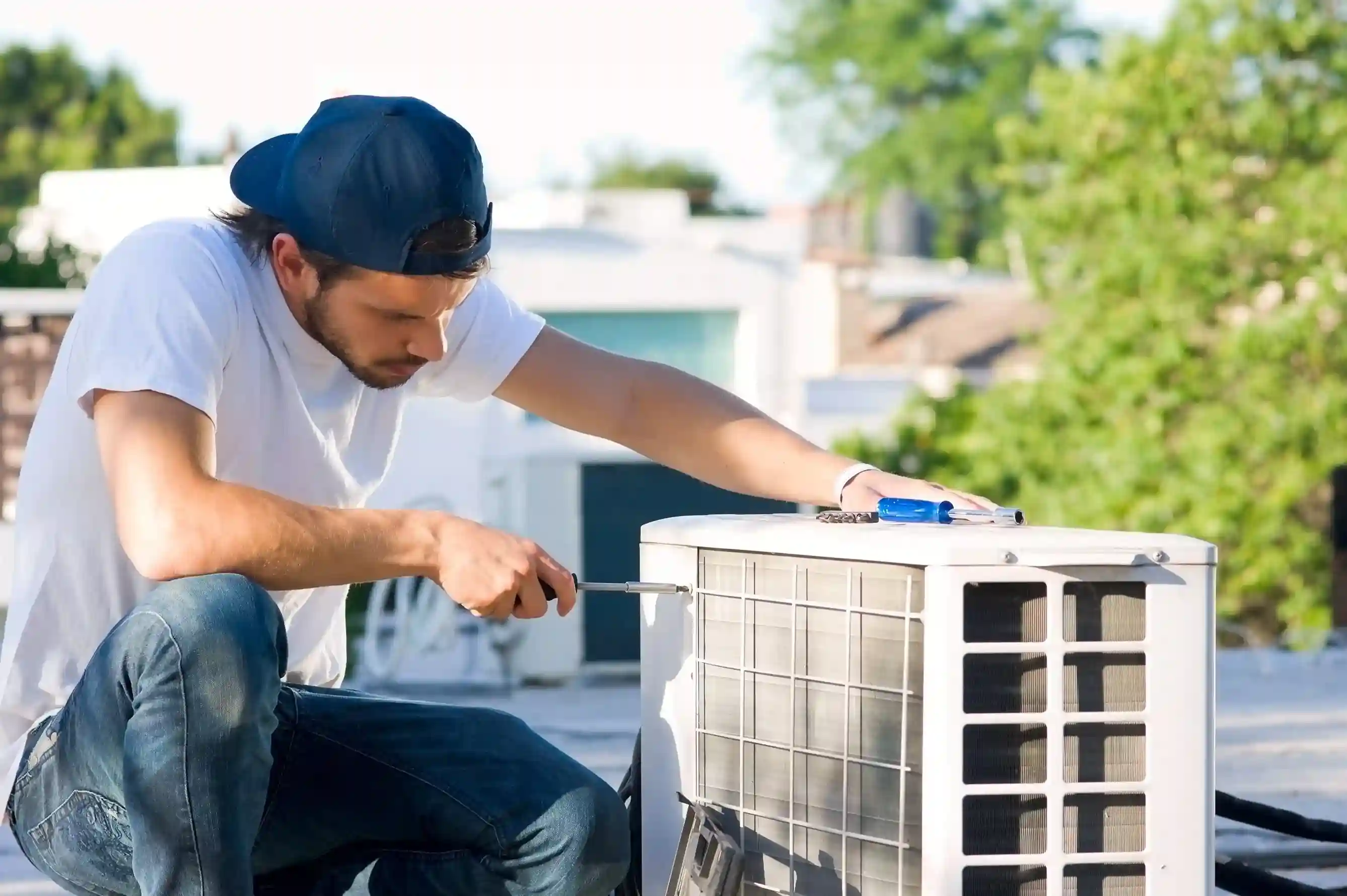 Technician in white shirt and blue cap servicing rooftop AC unit with screwdriver, cityscape in background