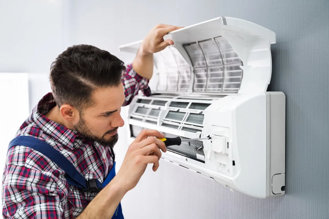 Person in plaid shirt using screwdriver to service wall-mounted air conditioner with open front panel