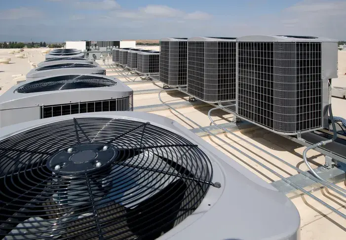 A vast white rooftop is filled with numerous commercial HVAC units, each with a large fan on top, extending into the distance under a blue sky.