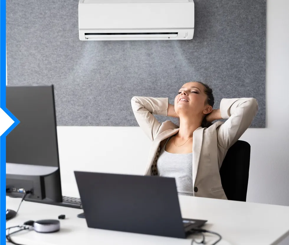 A relaxed woman is leaning back in her office chair at her desk, with a Daikin VRV air conditioner unit blowing cool air above her.