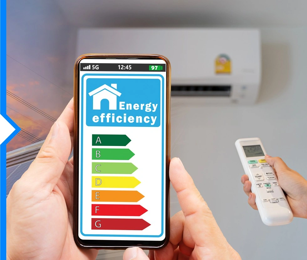 A person is holding a smartphone displaying an energy efficiency rating and a remote control for a wall-mounted air conditioner.