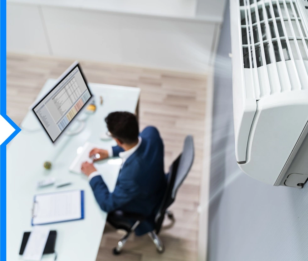 A high-angle view shows a man working at a desk while a wall-mounted air conditioning unit is blowing cool air.
