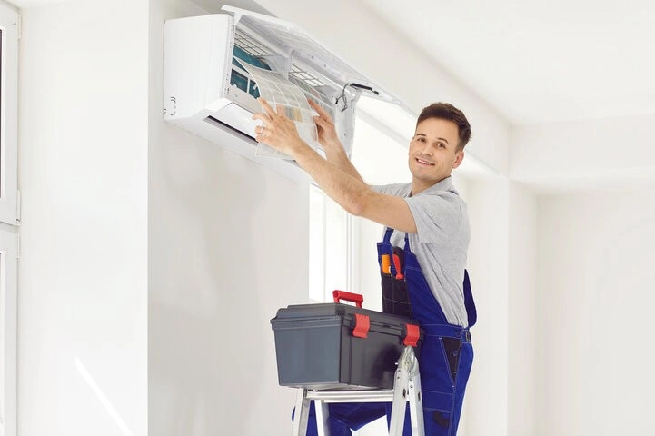 An HVAC technician on a ladder is smiling while servicing a Daikin air conditioner, removing the filter with a toolbox next to him.