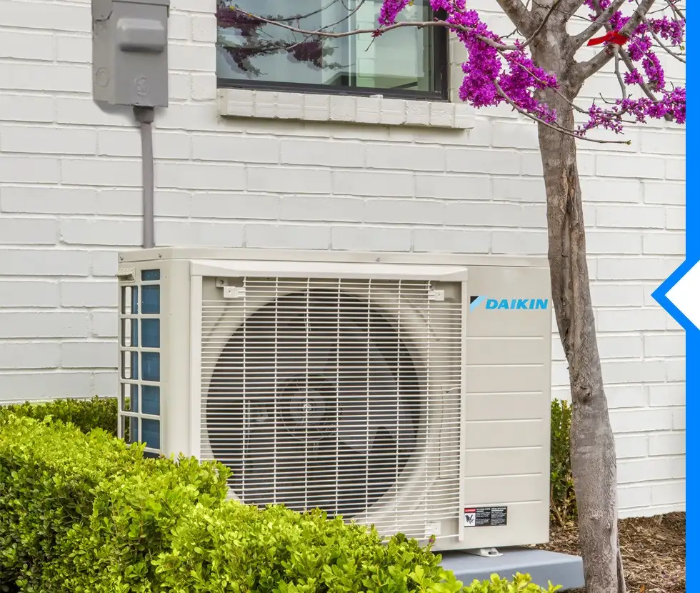 A Daikin FIT outdoor air conditioner is installed next to a white brick house, surrounded by green shrubs and a flowering tree.