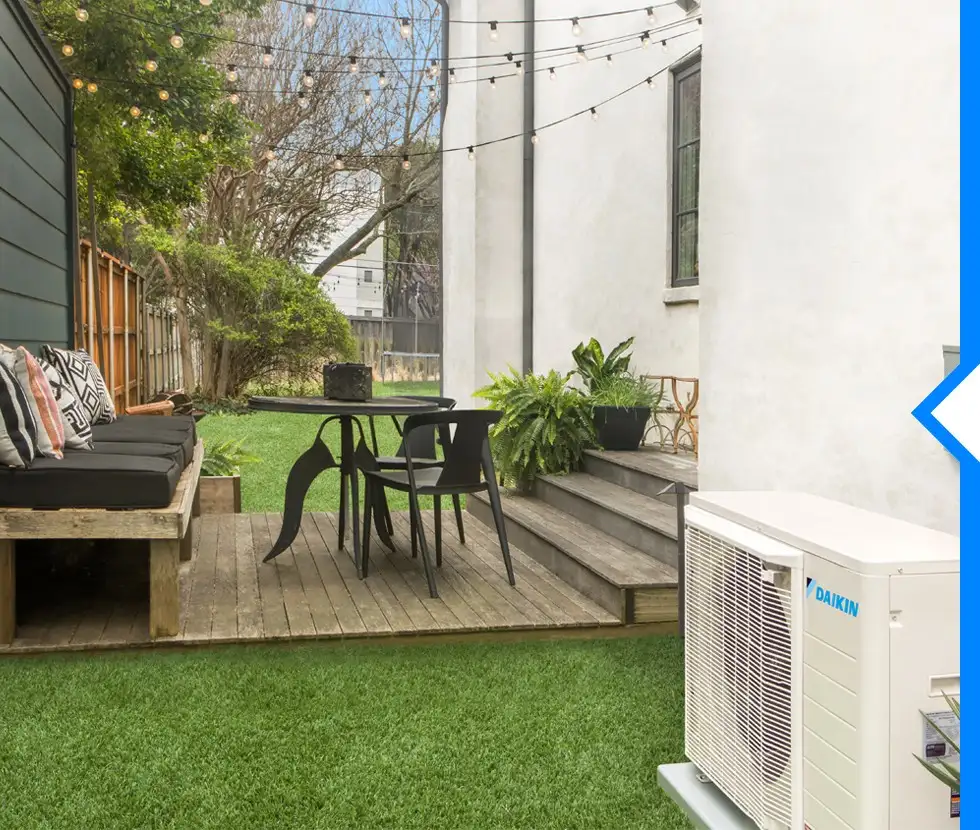 A Daikin FIT outdoor air conditioner unit is shown next to a wooden deck with a table and chairs, and a small backyard lawn.