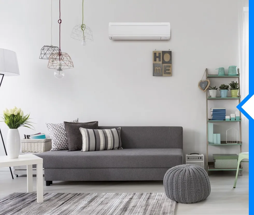 A white Daikin ductless AC unit is mounted on a white wall above a gray sofa with throw pillows and a shelf.