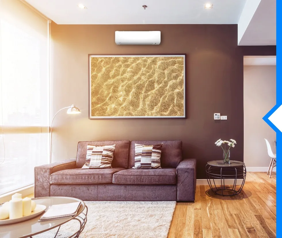 A white Daikin ductless AC unit is mounted on a dark brown wall above a sofa with a framed picture of sand.