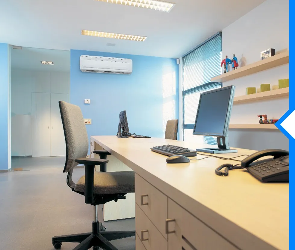 A Daikin ductless AC unit is mounted on a light blue wall in a modern office with desks, chairs, and computers.