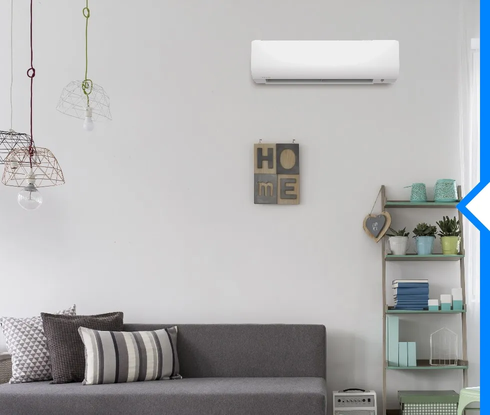 A white Daikin ductless AC unit is mounted on a white wall above a gray couch with throw pillows and a teal bookshelf.