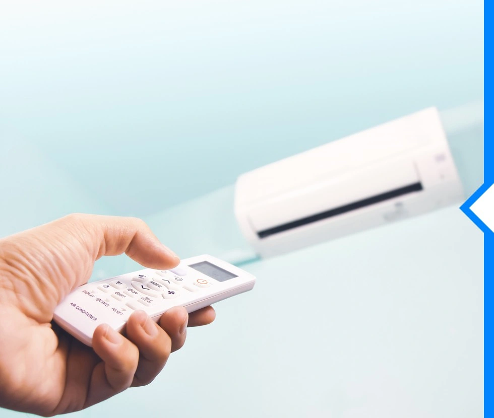 A hand is holding an air conditioner remote control, pointing it towards a blurry, wall-mounted white AC unit in the background.