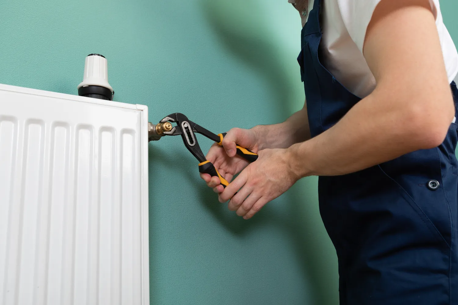 Plumber using pliers to repair radiator against teal wall