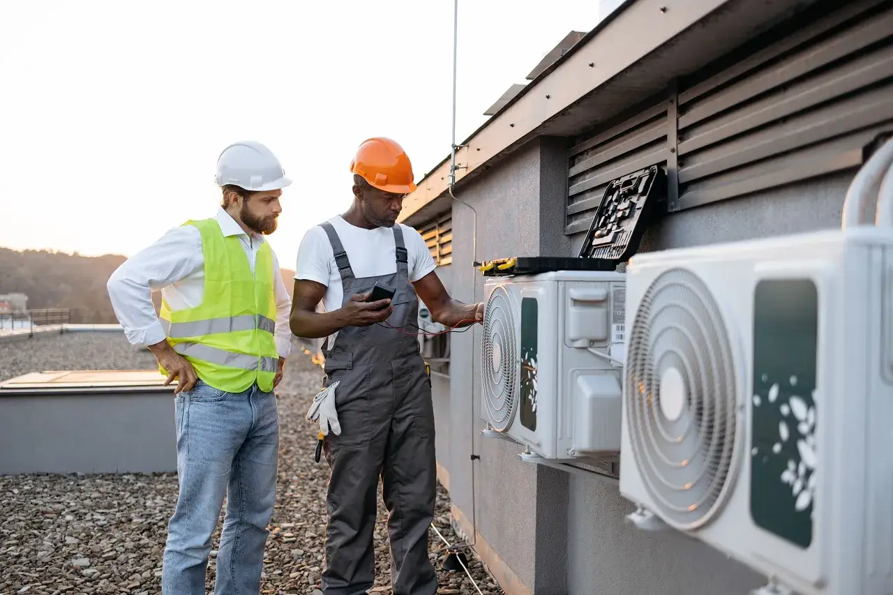 Two workers in safety helmets inspect air conditioning units on rooftop