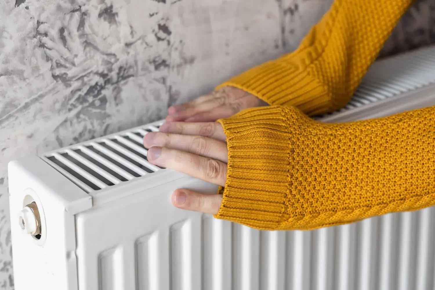 Hands in yellow sweater warming up on a white radiator heater