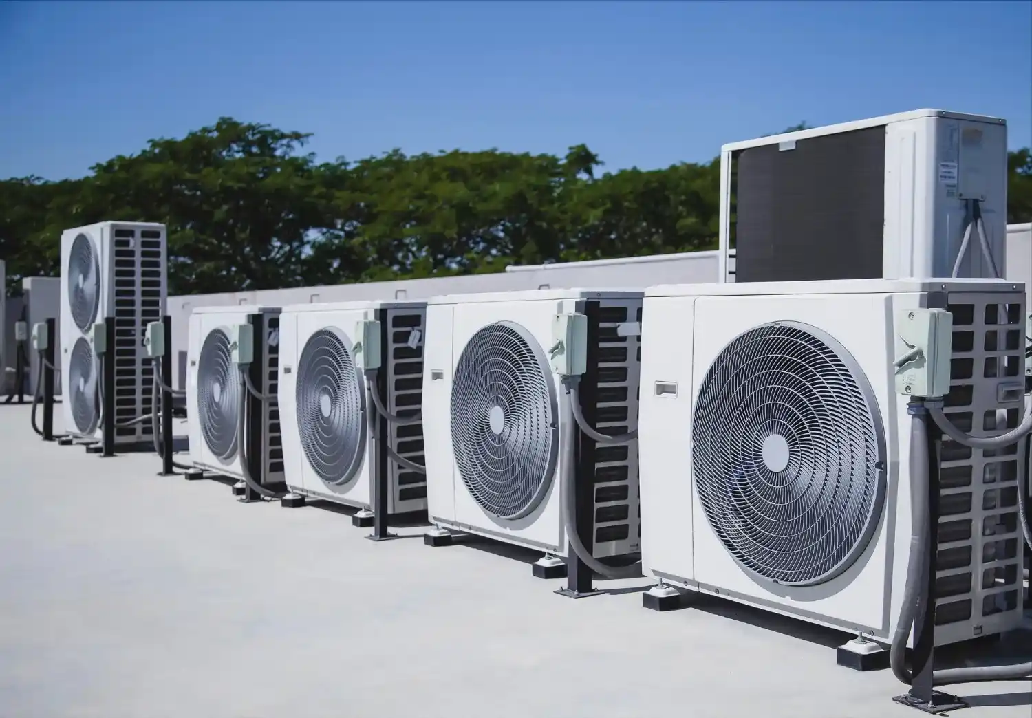 Multiple white air conditioning units lined up against blue sky and trees