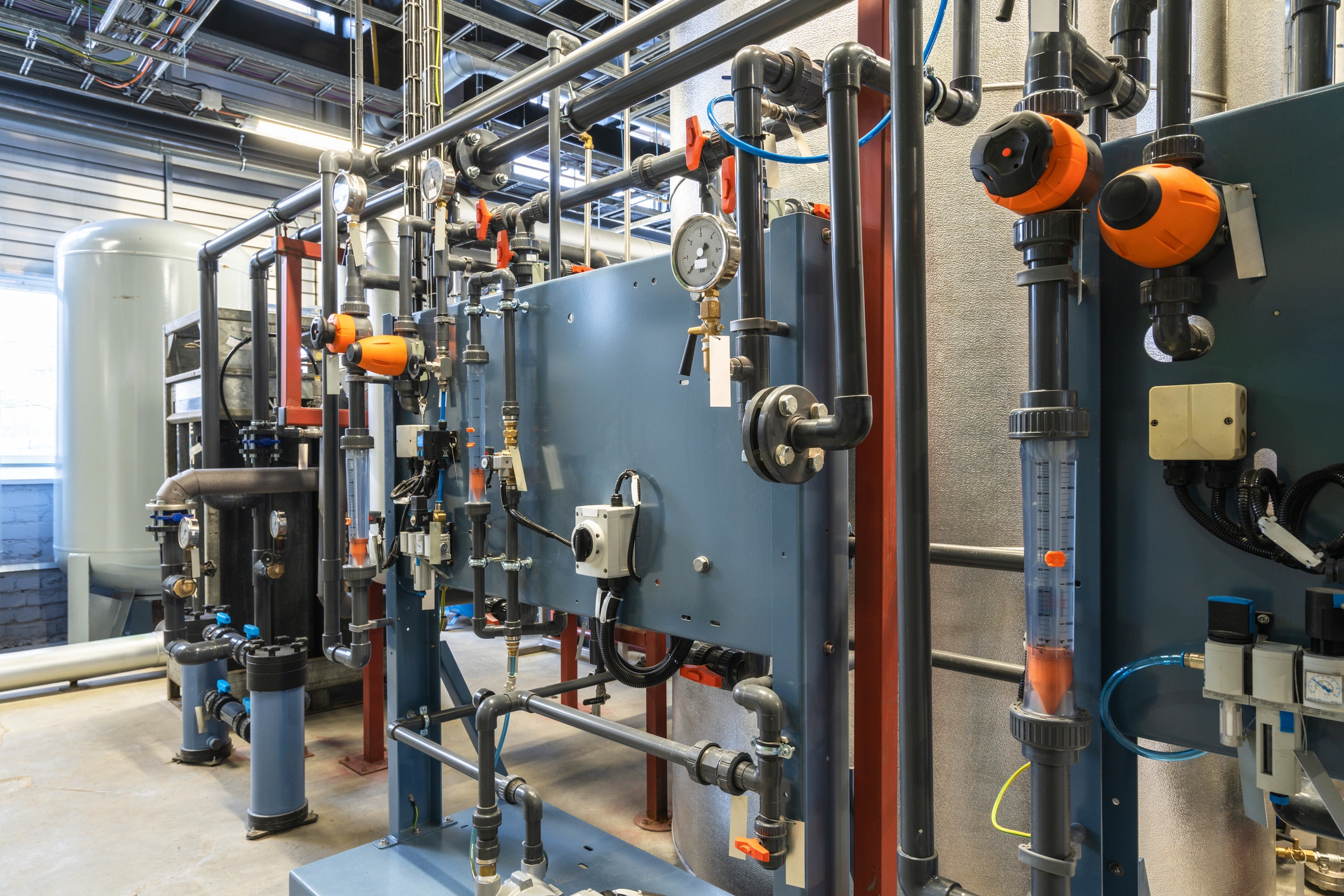 Industrial equipment room with intricate piping, gauges, and control panels. Orange safety features stand out.