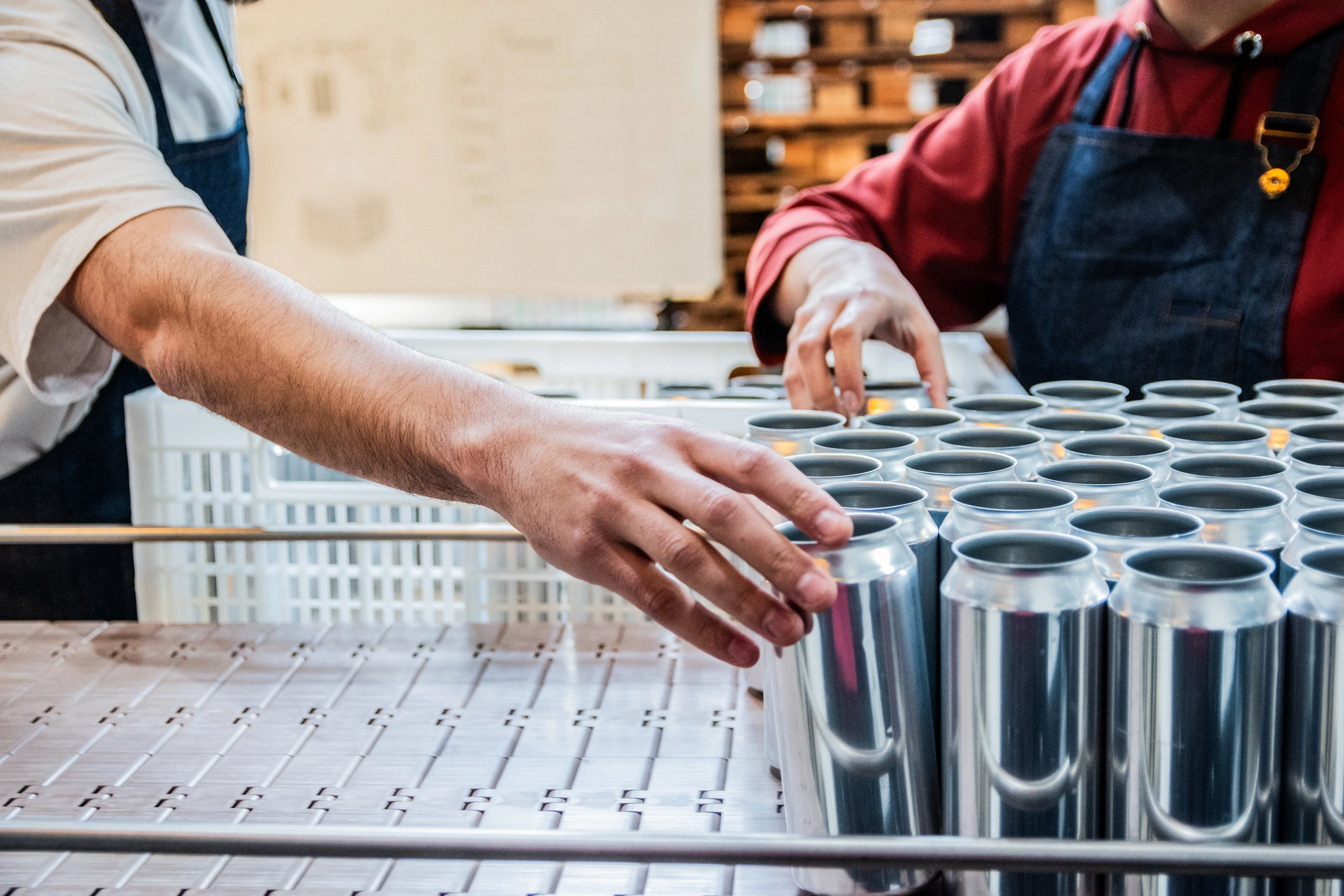 Two people in aprons work in a brewery, organizing rows of aluminum beer cans on a table.