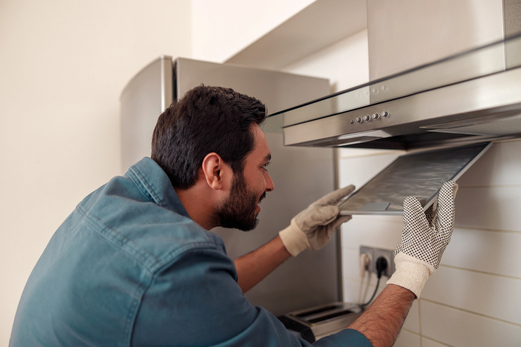 A man wearing a blue shirt and gloves repairs a stove, focused on fixing its components.