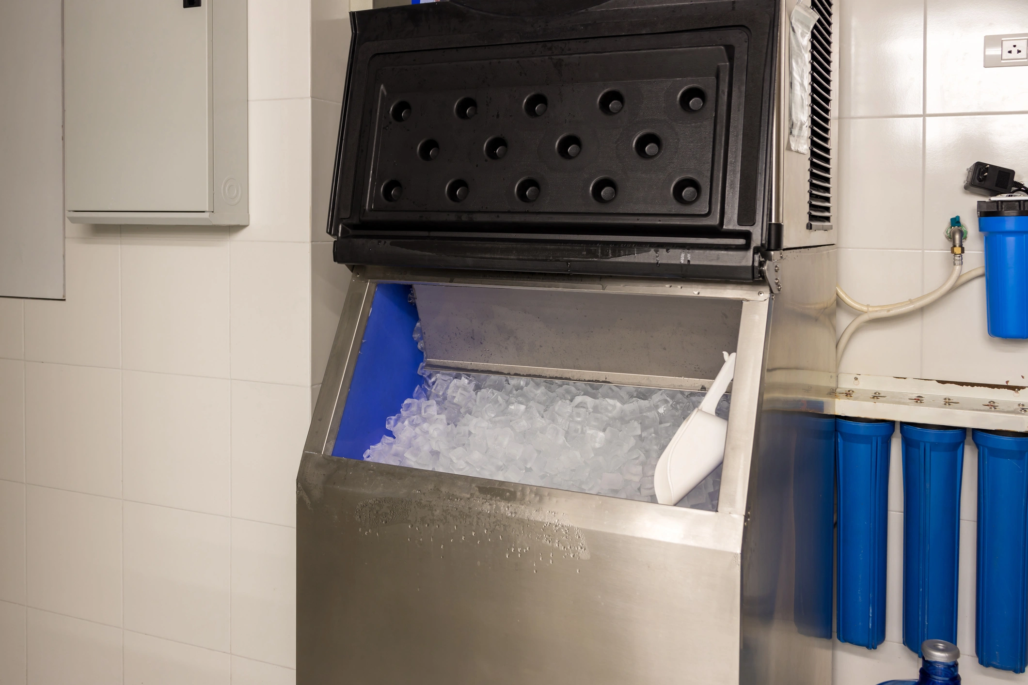 Commercial ice maker in a tiled room, filled with clear ice cubes and a white scoop.