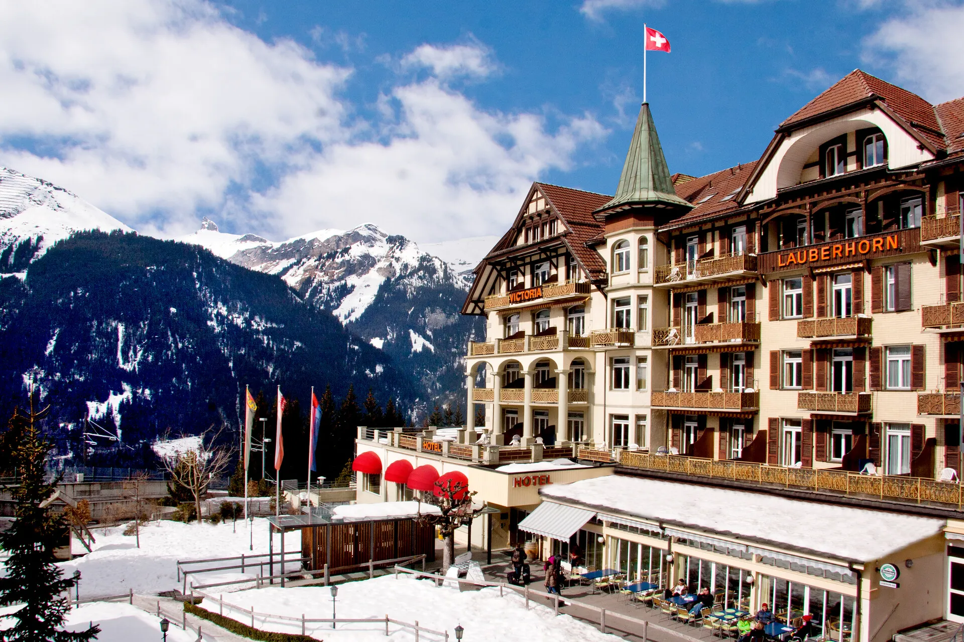 Snow-covered hotel with red awnings and Swiss flag against a backdrop of snowy mountains and blue sky.