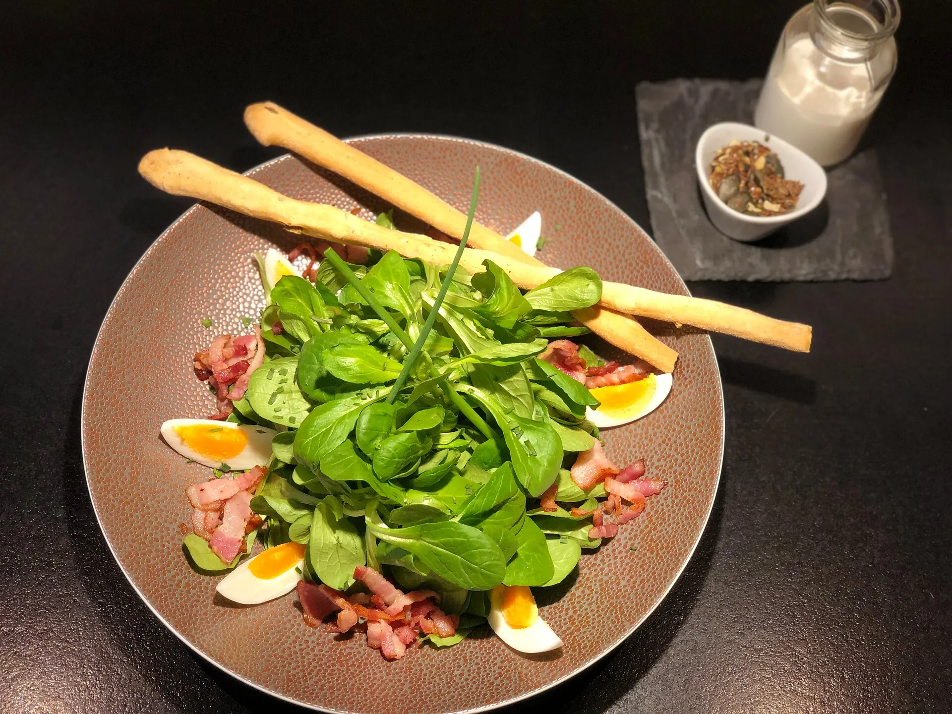 Salad with greens, boiled egg wedges, bacon, two breadsticks, seeds, and a jar of dressing on black surface.