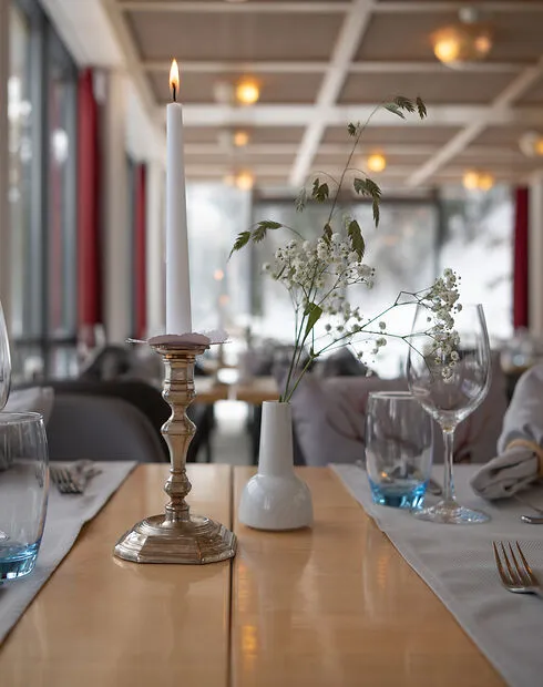 Table set with a lit candle in a brass holder and a small white vase with flowers in a restaurant.