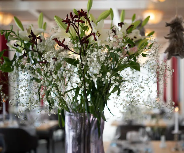 Vase of white lilies and small white baby's breath flowers on a table in a softly lit room.
