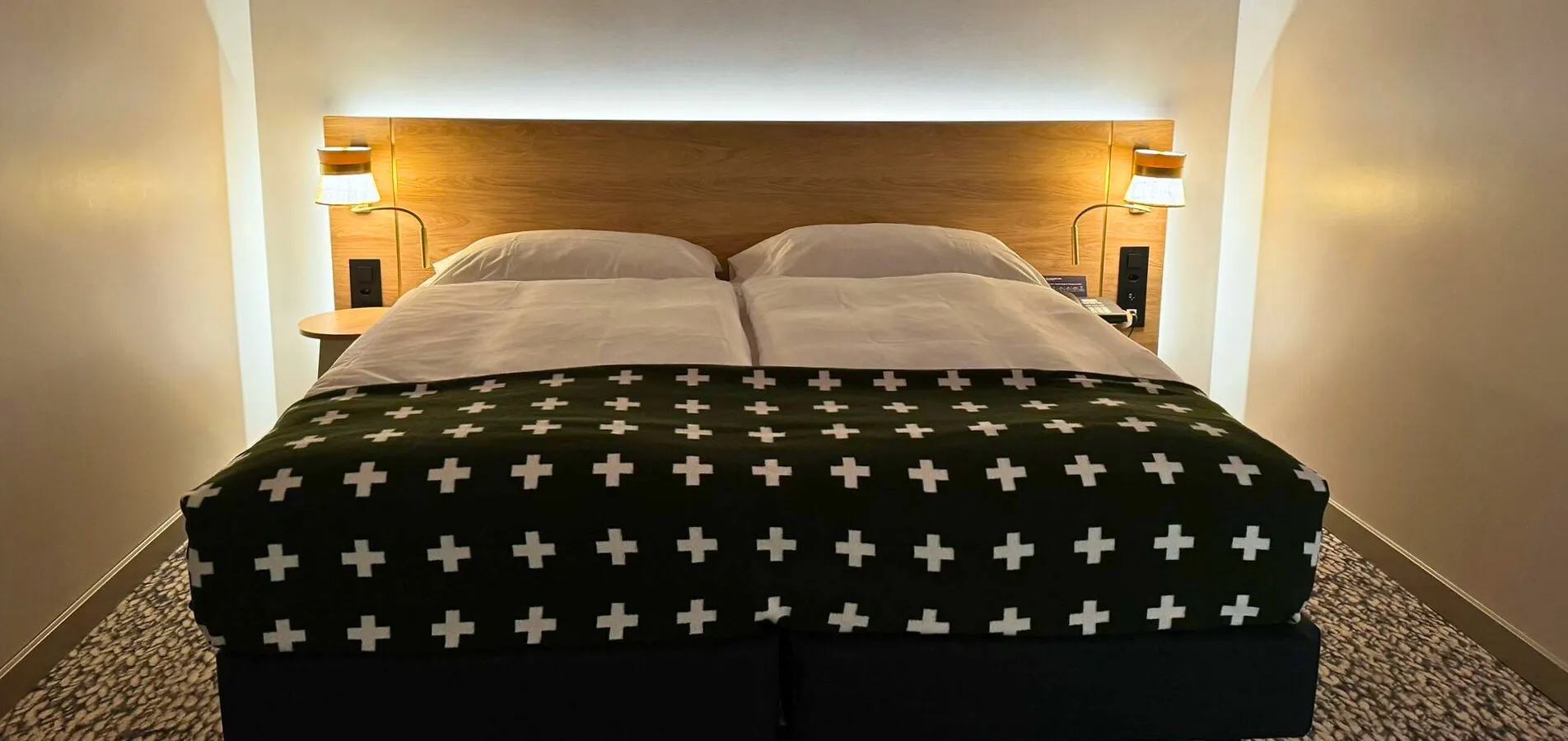 Double bed with white sheets and a black blanket with white crosses in a softly lit bedroom.