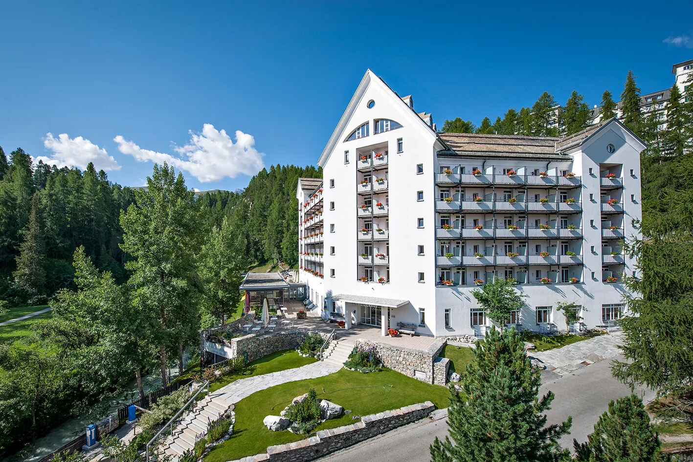 White multi-story hotel with balconies and flowers, surrounded by green trees under a blue sky.