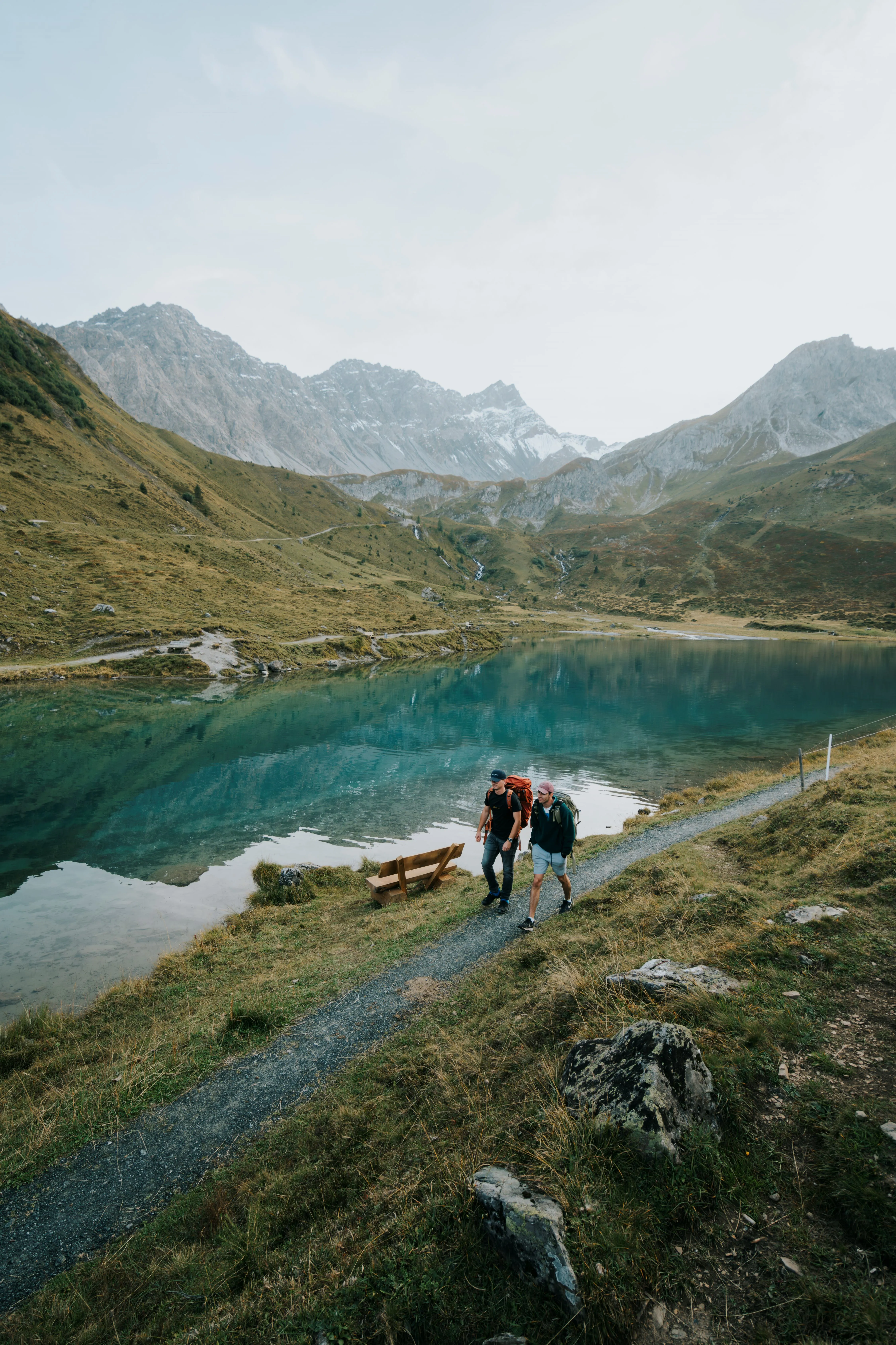 Two hikers walk on a path beside a clear blue lake surrounded by mountains.