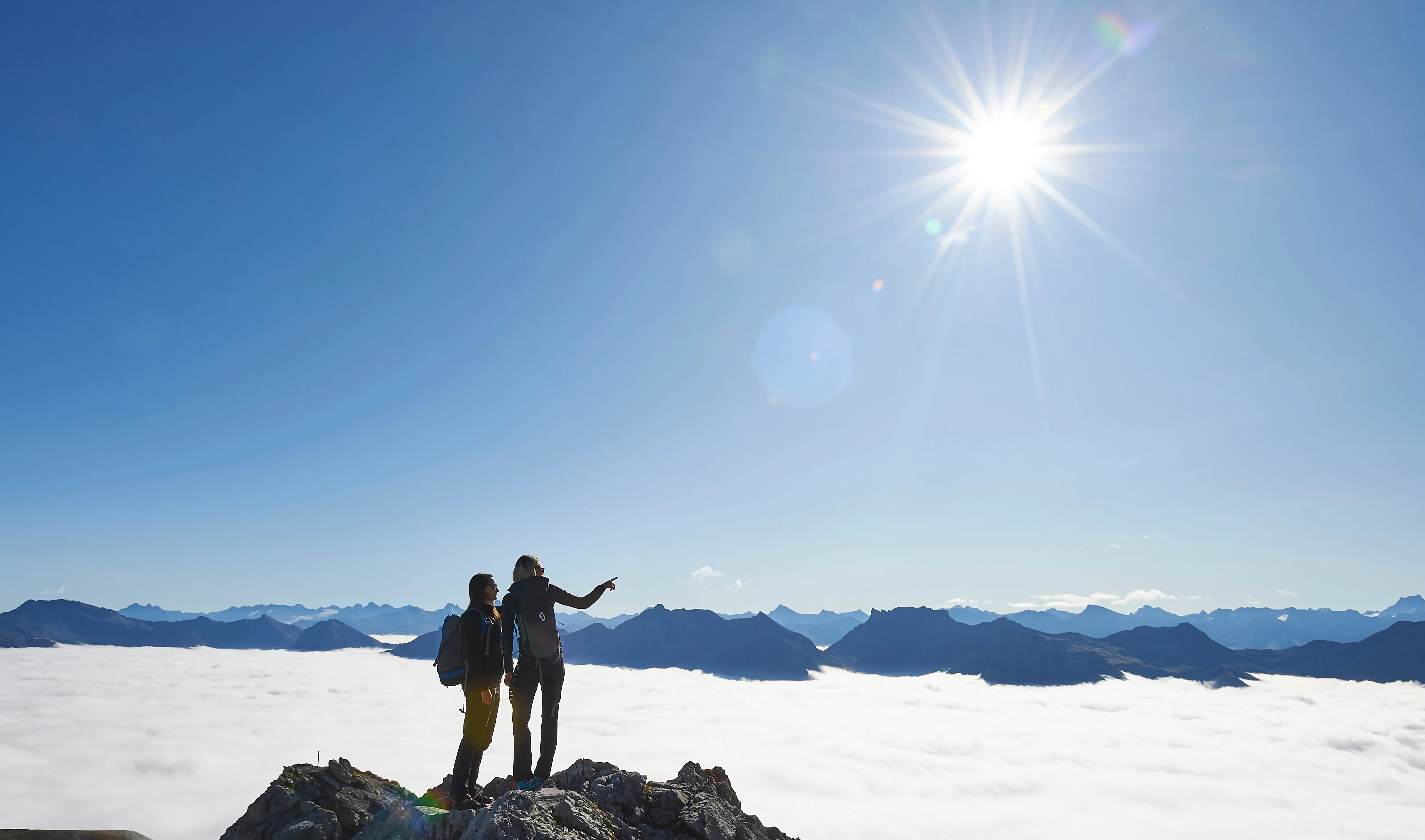 Two hikers with backpacks stand on rocky peak above a cloud-covered mountain range under bright sun.