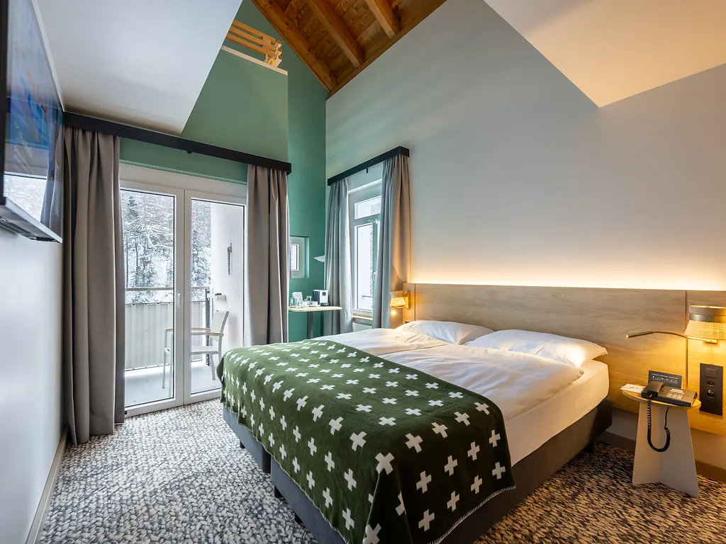 Modern hotel room with a double bed, green patterned blanket, wood ceiling, and balcony doors with curtains.