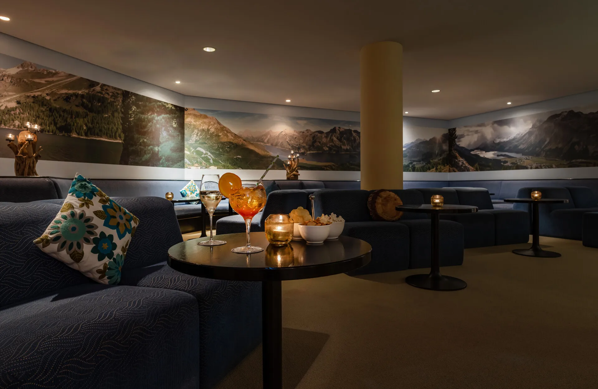 Cozy lounge with dark sofas, floral pillows, small tables, drinks, snacks, and panoramic mountain murals.