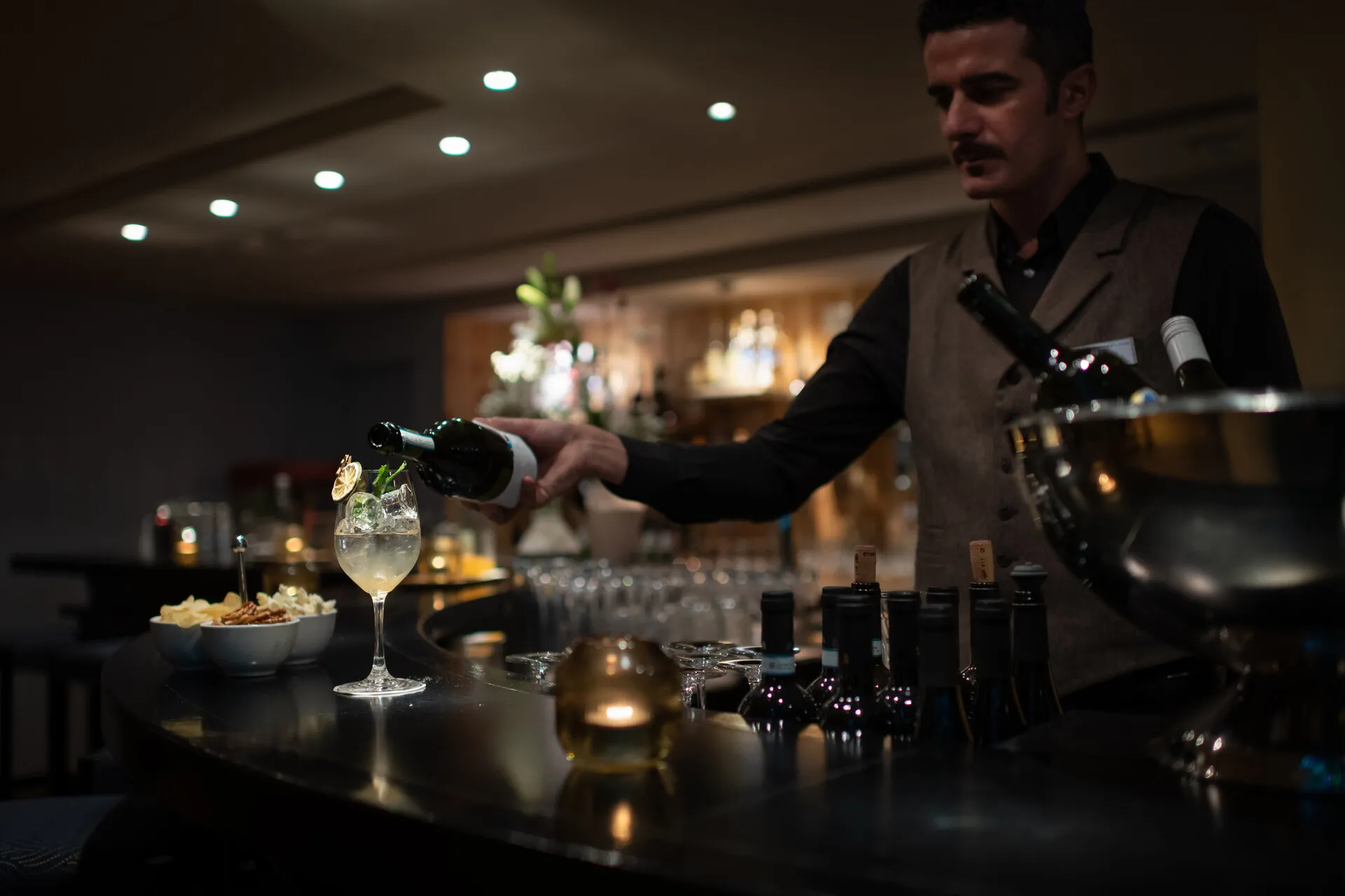 Bartender pouring a drink from a bottle into a glass with ice and garnish at a bar counter.
