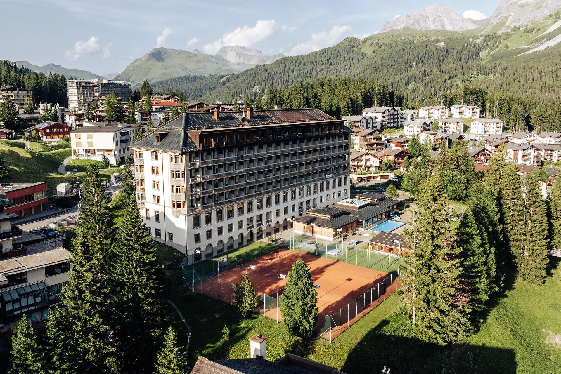 Hotel with tennis courts and pool surrounded by pine trees in a mountain village under blue sky.