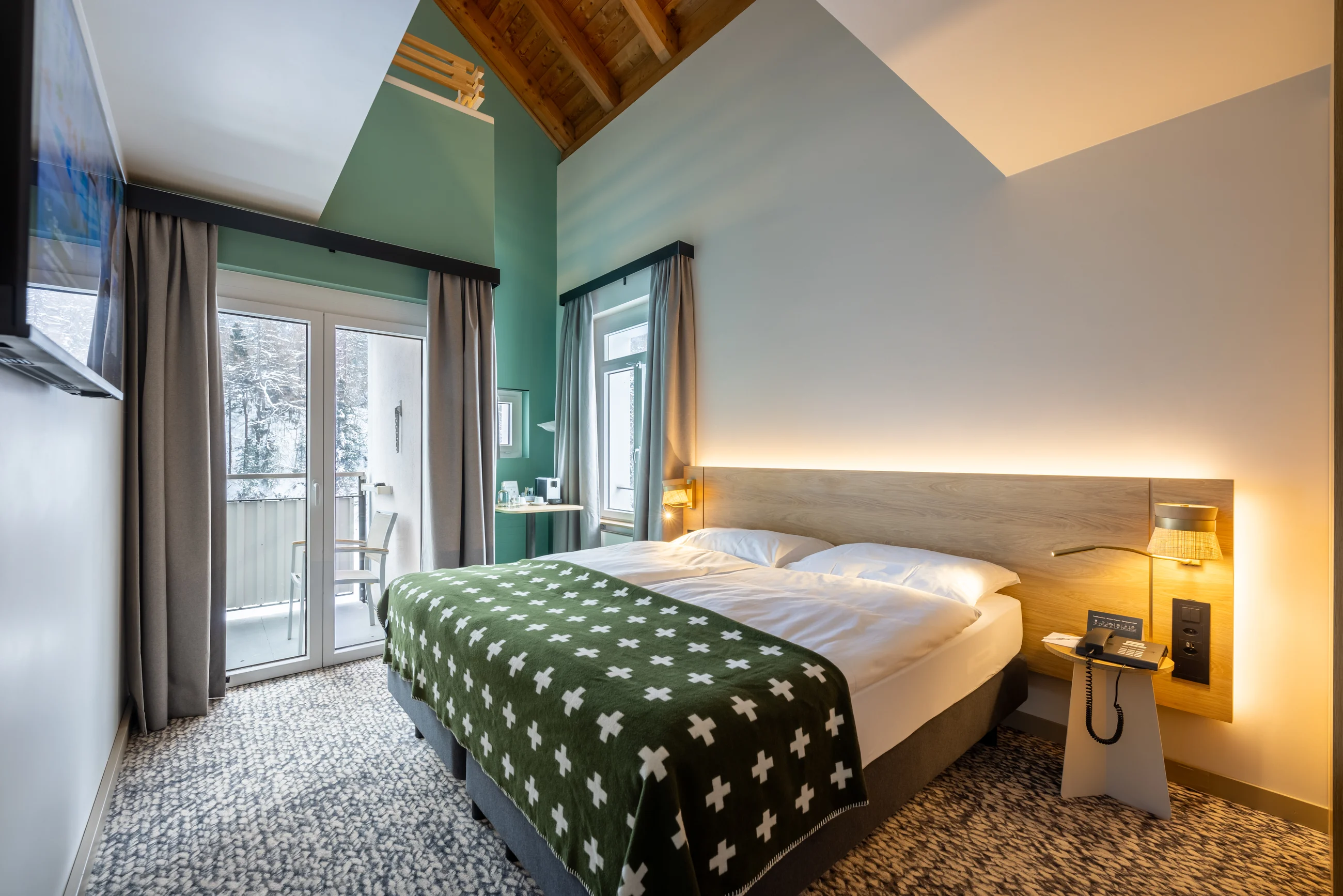 Modern hotel room with a double bed, green patterned blanket, wood ceiling, and balcony doors with curtains.