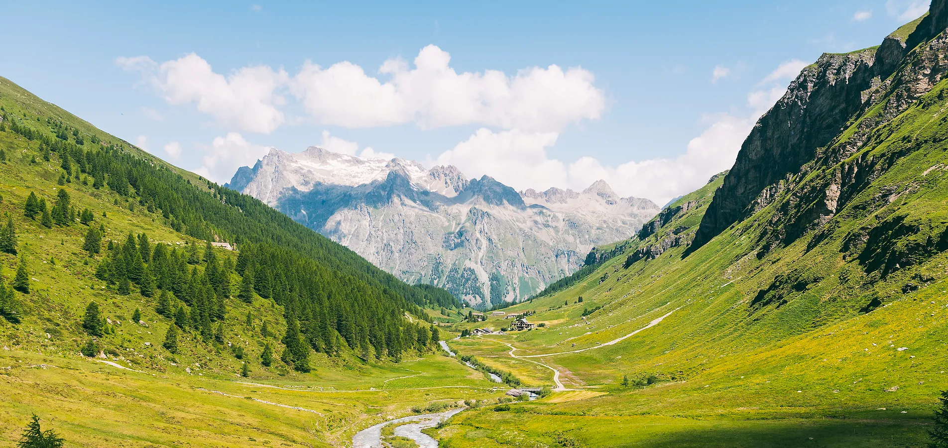 Ein üppig grünes Bergtal mit einem kleinen blauen See und verschlungenen Pfaden unter teilweise bewölktem Himmel.