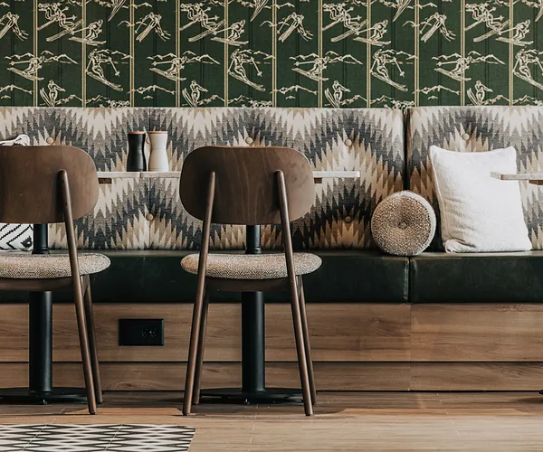 Modern café seating with patterned bench, two wood chairs, pillows, and wallpaper with skiing figures.