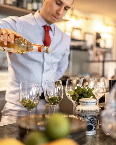 Man in red tie pouring liquid into wine glasses with mint leaves in a bar setting.