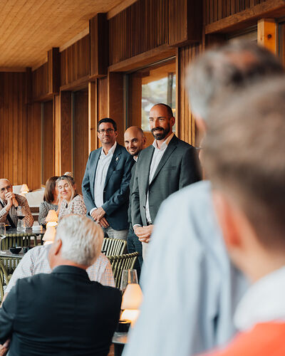 Three men in suits standing and smiling in a warmly lit wooden room with seated audience.