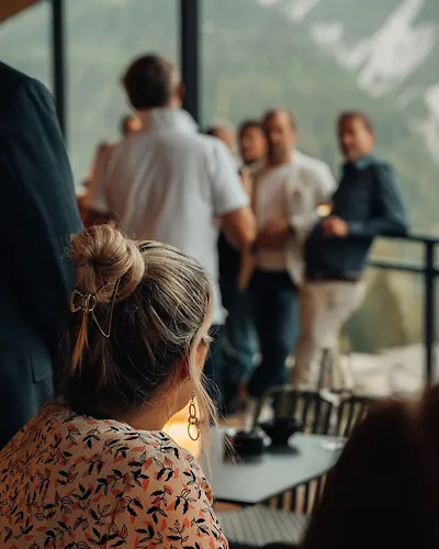 Woman with hair in a bun and floral top looking toward a group of people talking by a window.